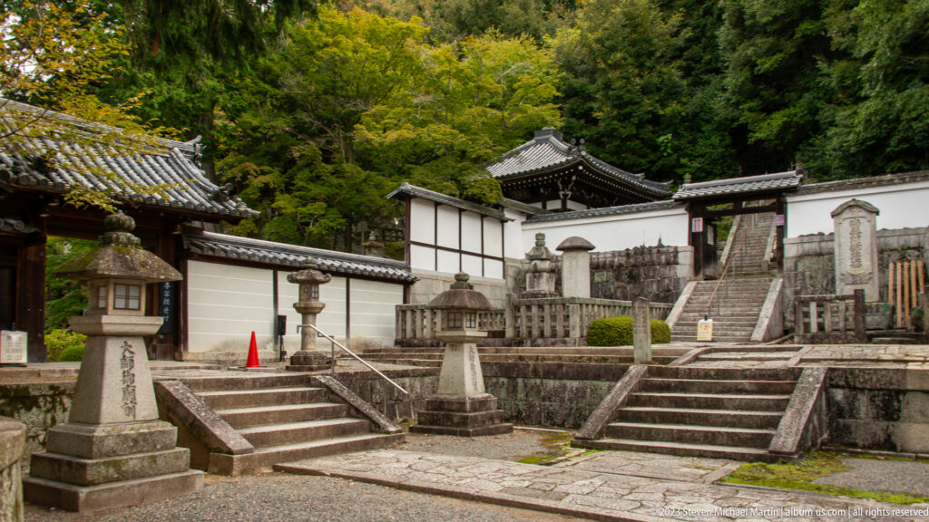 Terraced Buddhist cemetery with stone memorials, lanterns, steps, walls, and small temple buildings among trees.