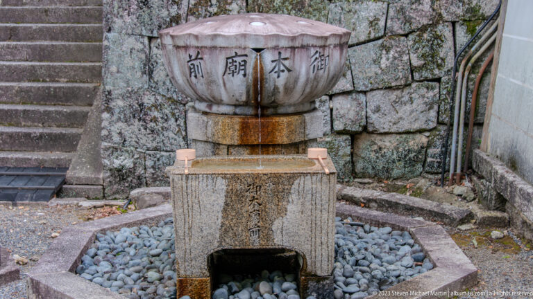 Inscribed stone water basin beside a masonry wall and steps, with Japanese characters around the upper bowl.
