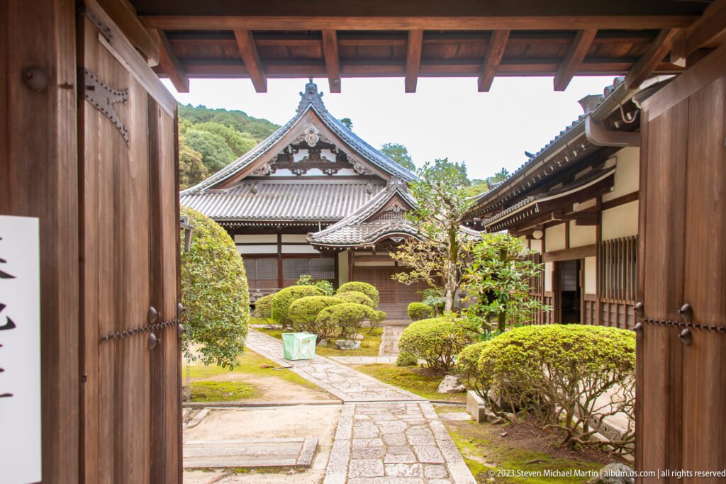 Open wooden gate framing a small garden with clipped shrubs, stone paving, and tiled temple buildings.
