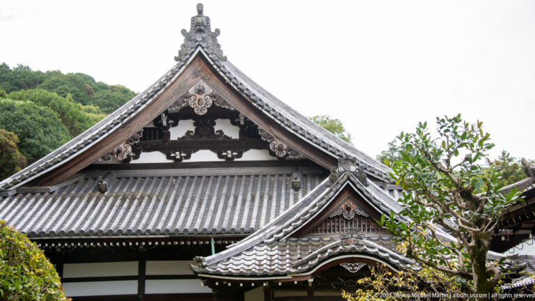 Ornate triangular temple gable above layered gray tiled roofs and white walls, with carved decorative details.