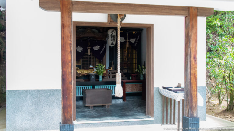 Open small shrine room with hanging bell rope, dark altar, flowers, ritual objects, and a Fudō Myōō plaque.