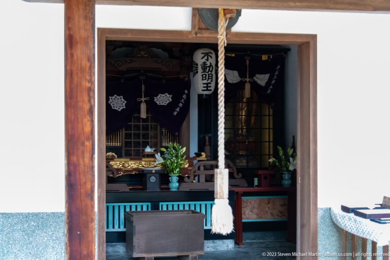 Close view into a Fudō Myōō altar chamber with bell rope, flowers, lamps, vessels, and dark curtains.