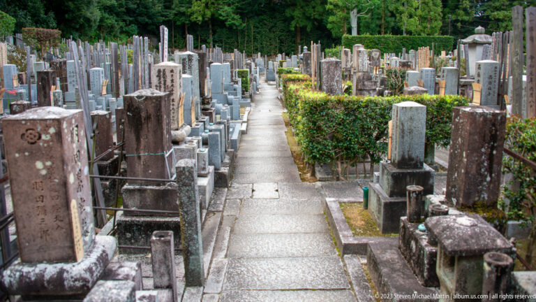 Narrow paved cemetery path between dense rows of weathered upright stone grave markers and clipped hedges.