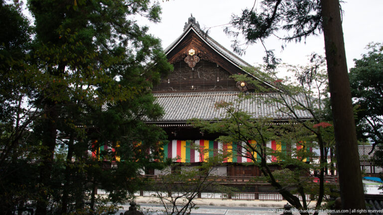 Dark timber temple hall seen through trees, with a tiled roof and five-colored Buddhist hangings along the façade.