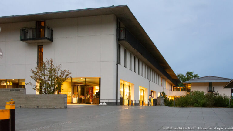 Modern white building with broad eaves, lit glass entrances, and a paved forecourt within the temple precinct.