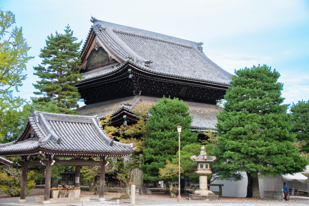 Large dark timber temple hall with massive tiled roof behind a smaller pavilion, pine trees, and stone lantern.