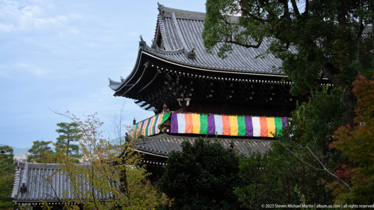 Dark timber temple structure partly hidden by trees, with five-colored Buddhist cloths beneath a steep tiled roof.