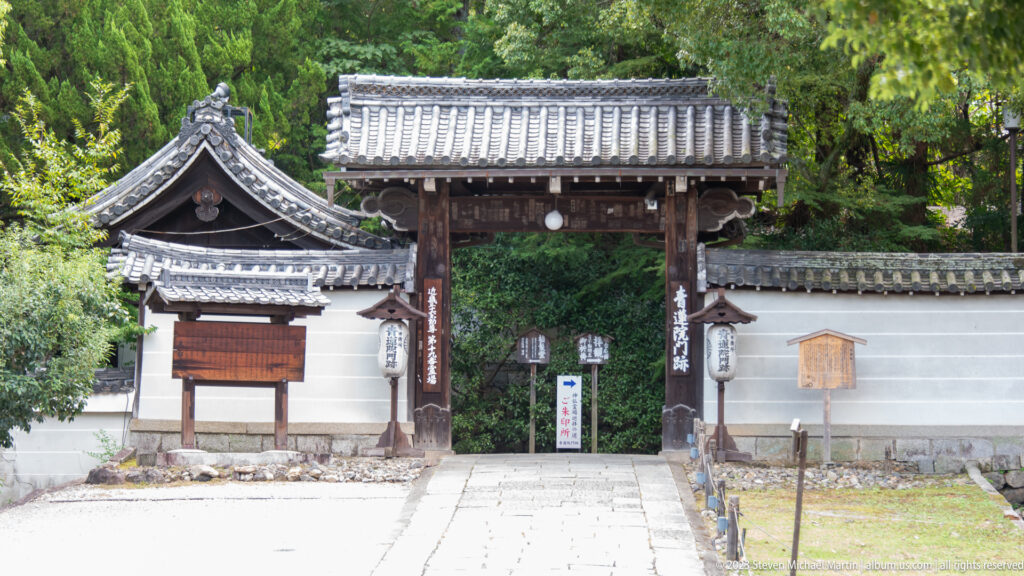 Small wooden gate in a white plaster wall with hanging lanterns, plaques, tiled roofs, and a path into greenery.