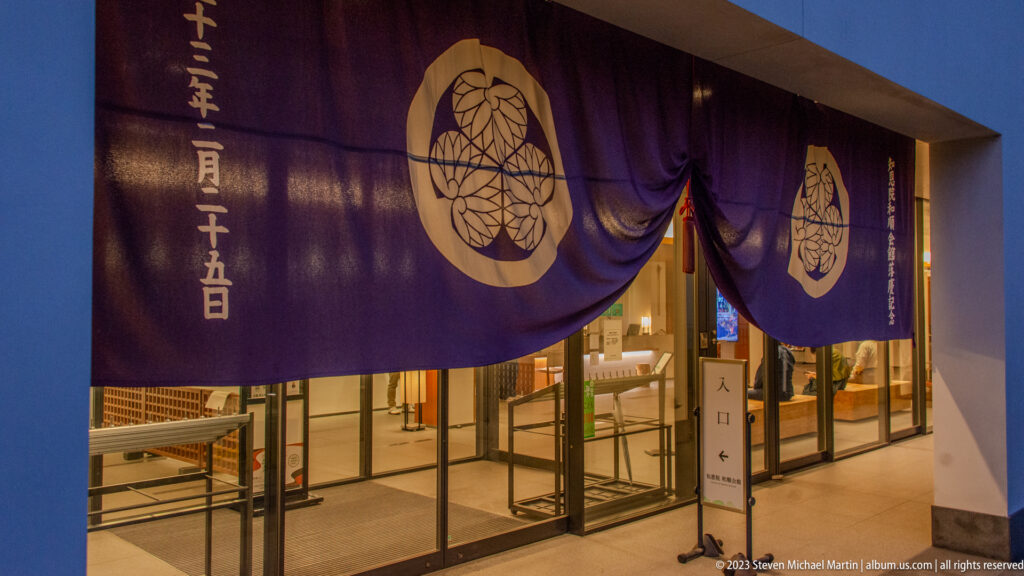 Purple temple curtain with repeated white hollyhock crests and vertical Japanese writing above a glass entrance.