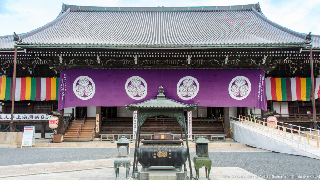 Frontal temple hall with purple crested curtains, multicolored hangings, steps, and a bronze incense burner centered in front.