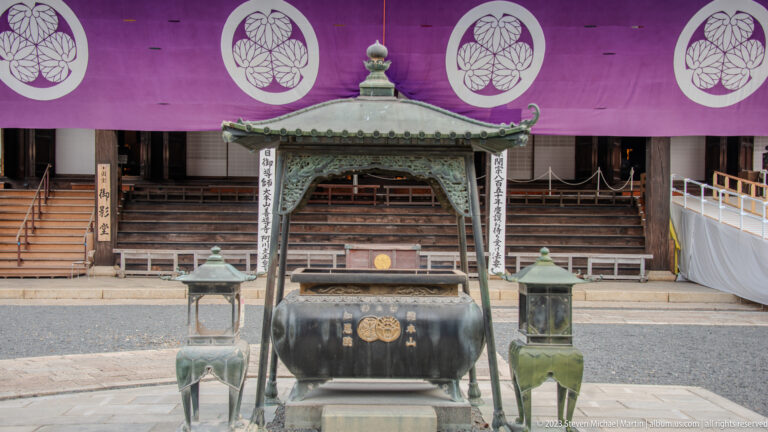 Large bronze incense burner between two metal lanterns, aligned with temple steps and purple curtains.