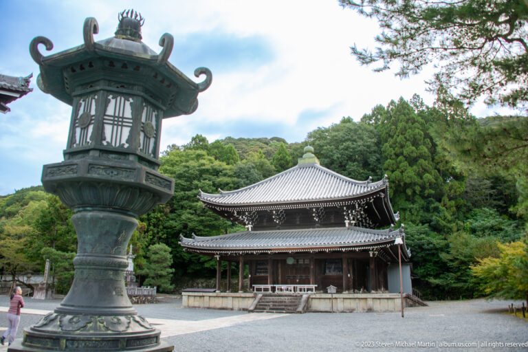Tall bronze temple lantern in the foreground with a dark wooden hall and forest behind.
