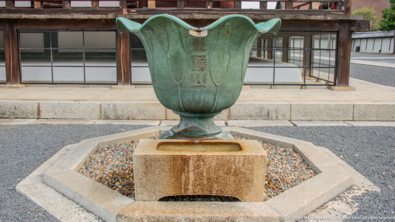 Green-patinated lotus-shaped metal basin on a square stone pedestal with a small Japanese inscription.