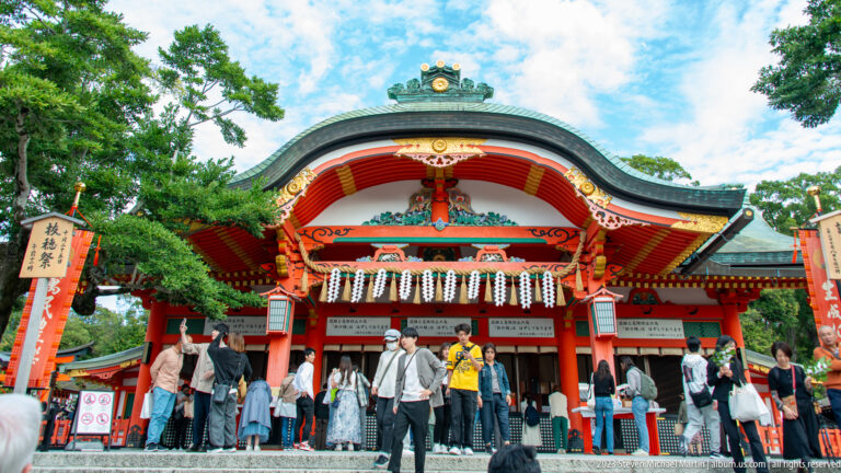 Crowded shrine entrance beneath a curved gable with painted carving, gold details, and white paper streamers.