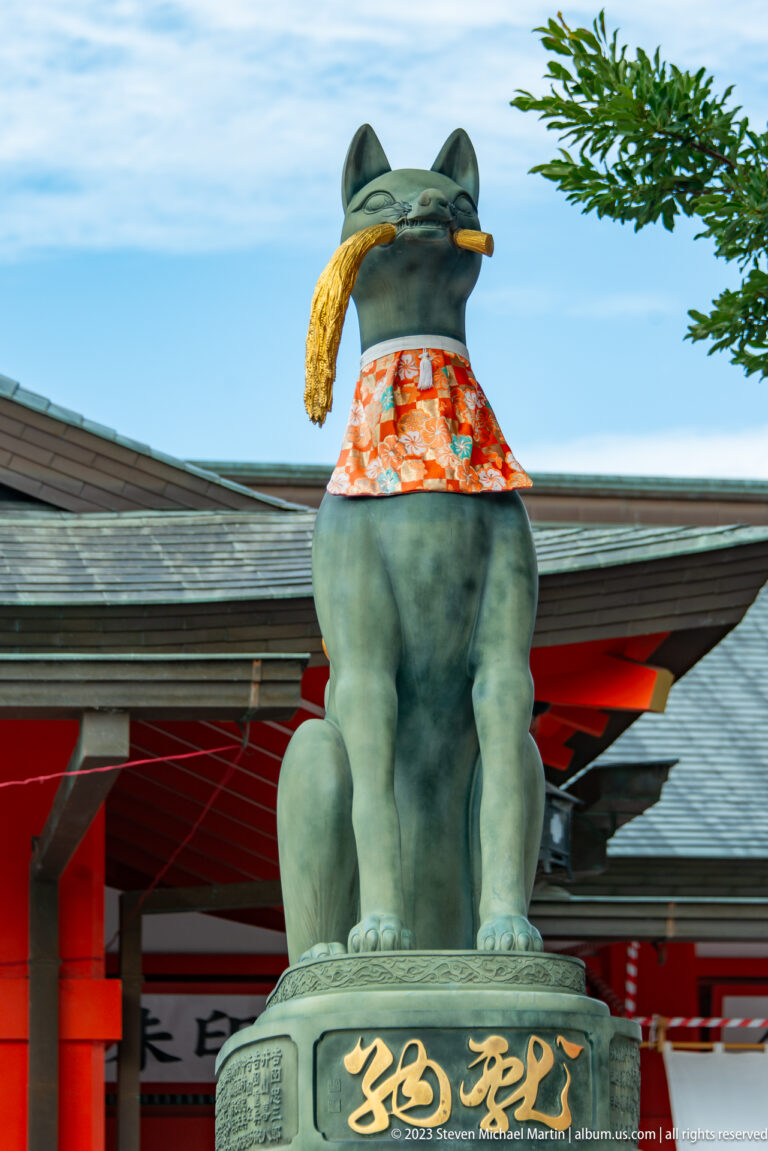 Seated fox guardian holding a golden rice sheaf, wearing a patterned red bib above an inscribed pedestal.