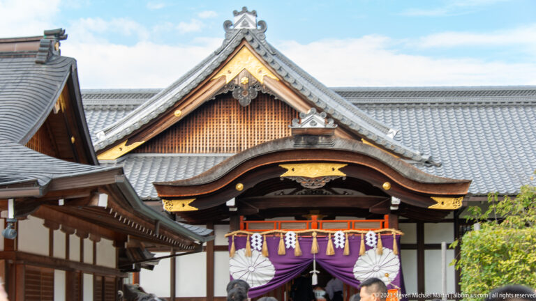 Curved shrine entrance with purple curtains, white paper streamers, and a larger tiled roof behind.