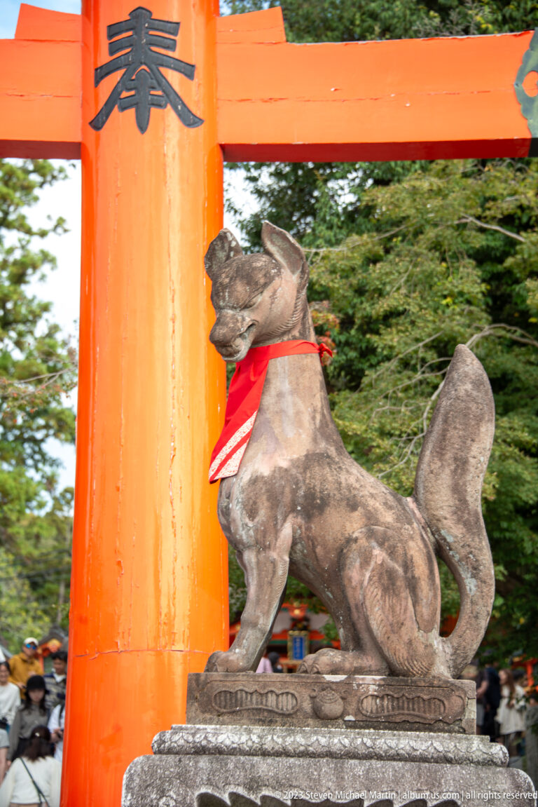 Weathered stone fox guardian in a red bib seated beside a large vermilion torii.