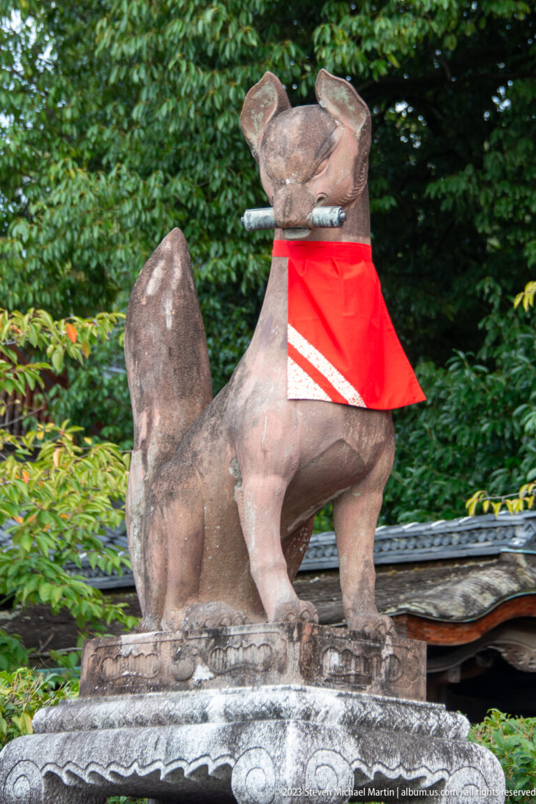 Front-facing stone fox guardian wearing a red bib on a carved pedestal against green foliage.