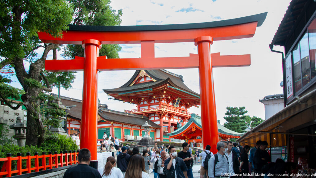 Crowds walking beneath a vermilion torii toward the two-story Rōmon gate at Fushimi Inari Taisha.