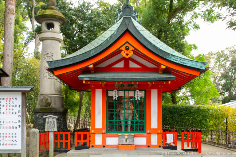 Small vermilion shrine with green roof beside a tall stone monument.