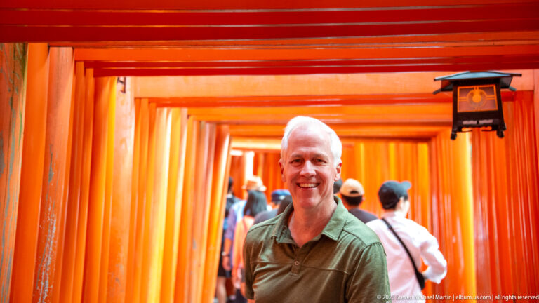 Visitor posing in the illuminated vermilion torii tunnel with pilgrims behind.