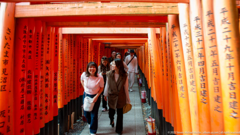Visitors walking through a narrow corridor of vermilion torii with donor inscriptions.