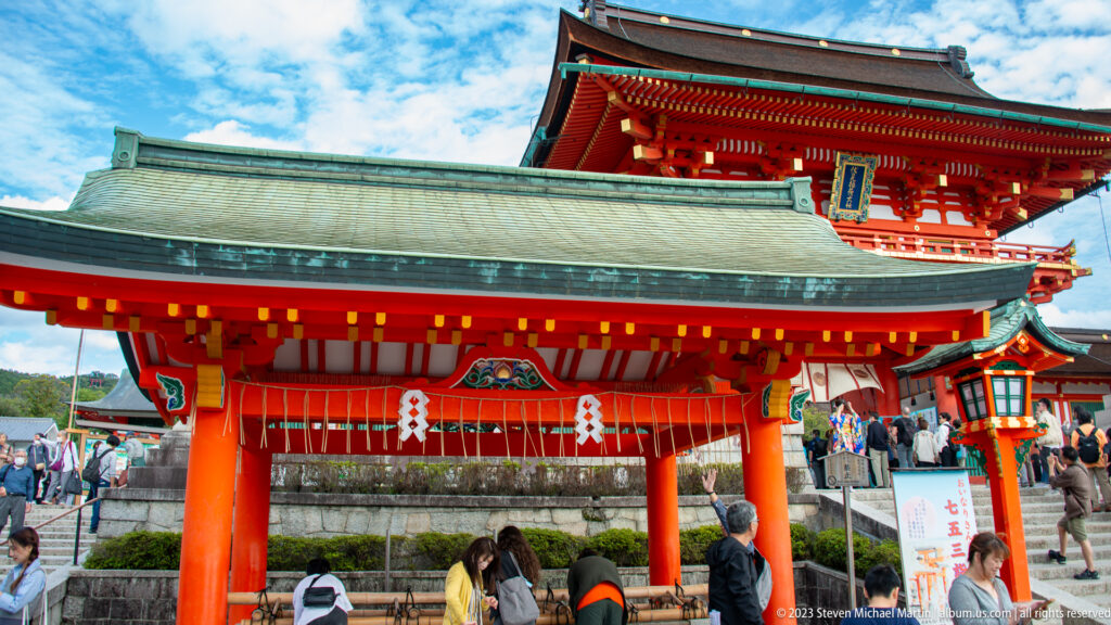 Roofed vermilion purification pavilion with stone basin, set before the taller Rōmon gate at Fushimi Inari Taisha.