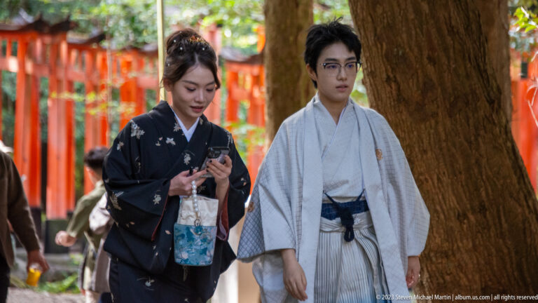 Two visitors in traditional kimono walking beneath trees with vermilion torii behind them.