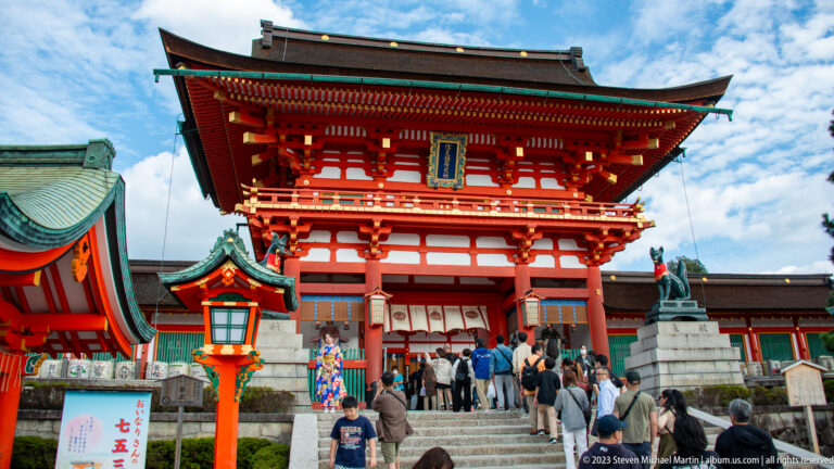 Frontal view of the vermilion Rōmon gate above stone steps, with visitors and a fox statue at right.