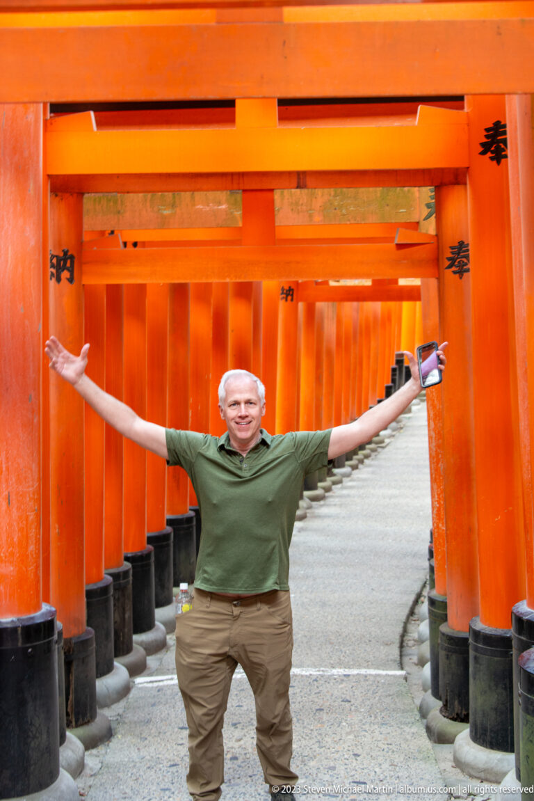 Visitor with outstretched arms standing inside a corridor of vermilion torii.