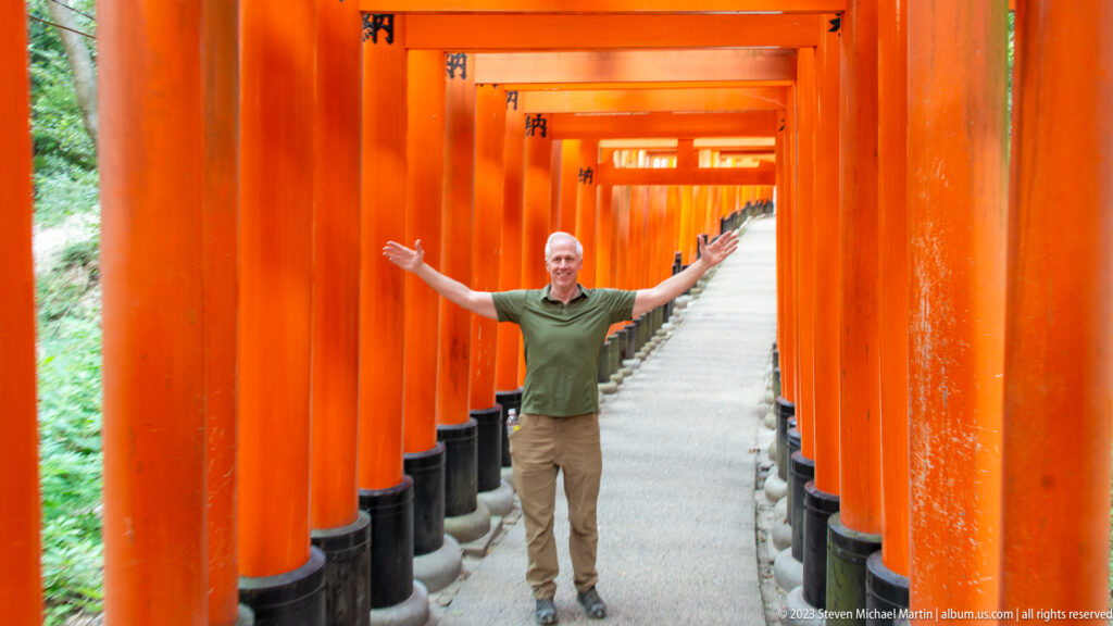 Visitor with arms extended on a rising path inside a corridor of closely spaced vermilion torii.