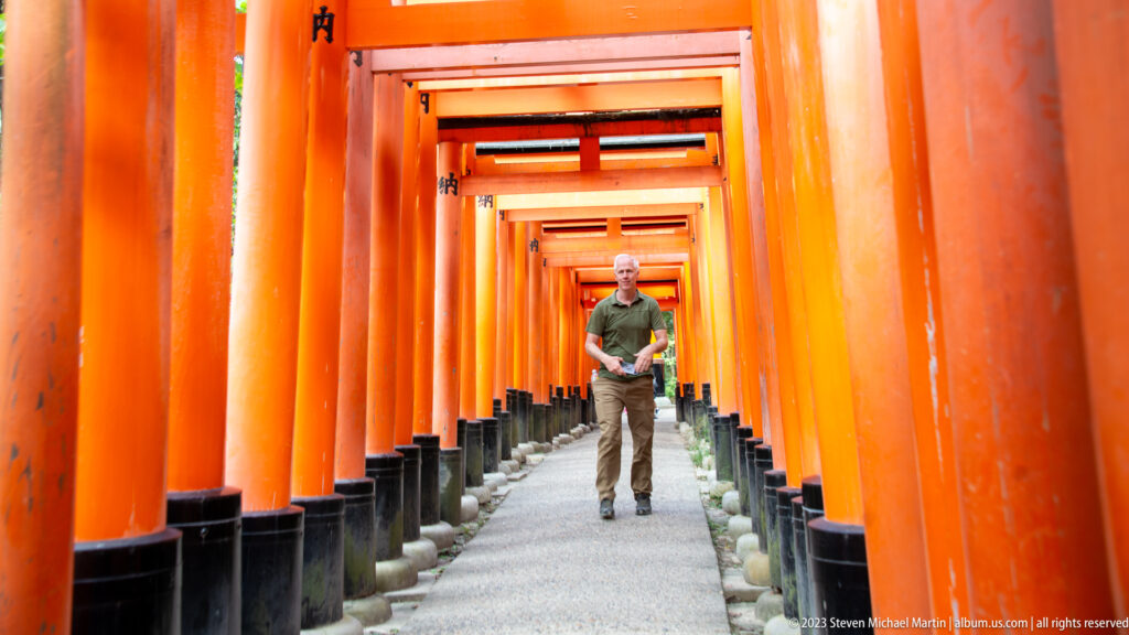 Visitor walking toward the camera along a gravel path enclosed by rows of vermilion torii.