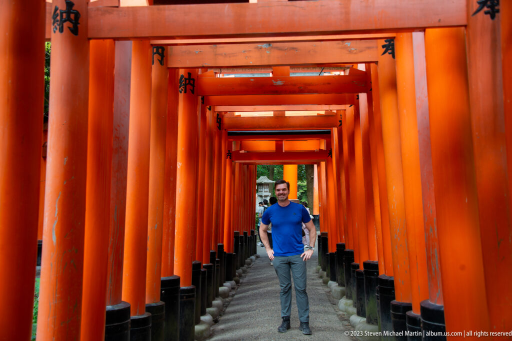 Full-length portrait of a visitor in a blue shirt centered inside a vermilion torii corridor.