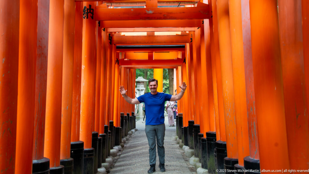 Visitor in a blue shirt raising both arms inside a vermilion torii corridor with people behind.