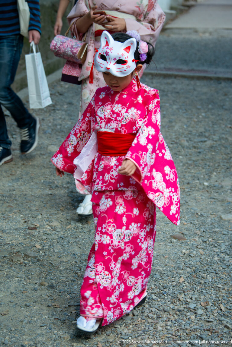 Child in a pink floral kimono walking on gravel while wearing a decorated white fox mask.