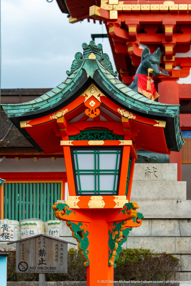Painted wooden lantern with white panels hanging beneath green, orange, and vermilion shrine eaves.