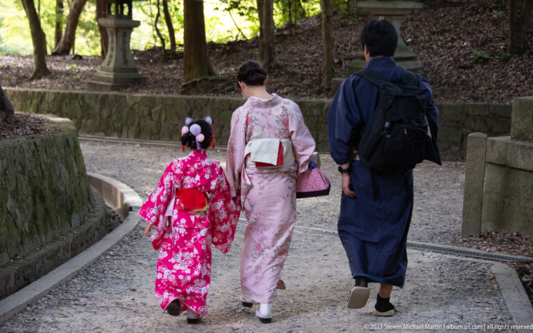 Adult and child in colorful kimono walking along a forested gravel shrine path.