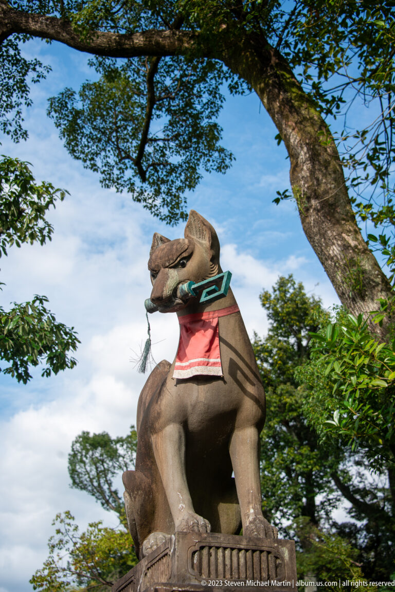 Stone fox guardian wearing a red votive bib beneath tree branches.
