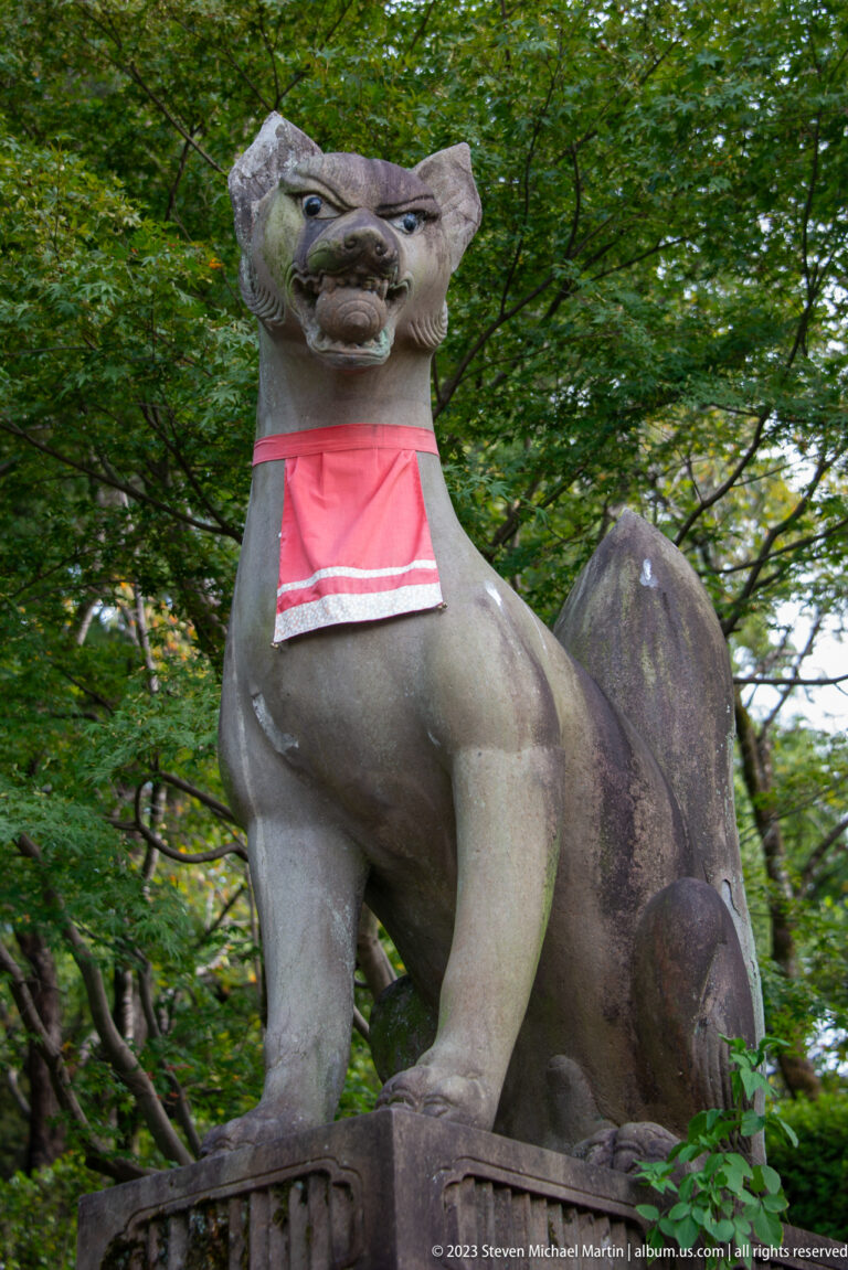 Front-facing stone fox guardian wearing a red votive bib before green foliage.