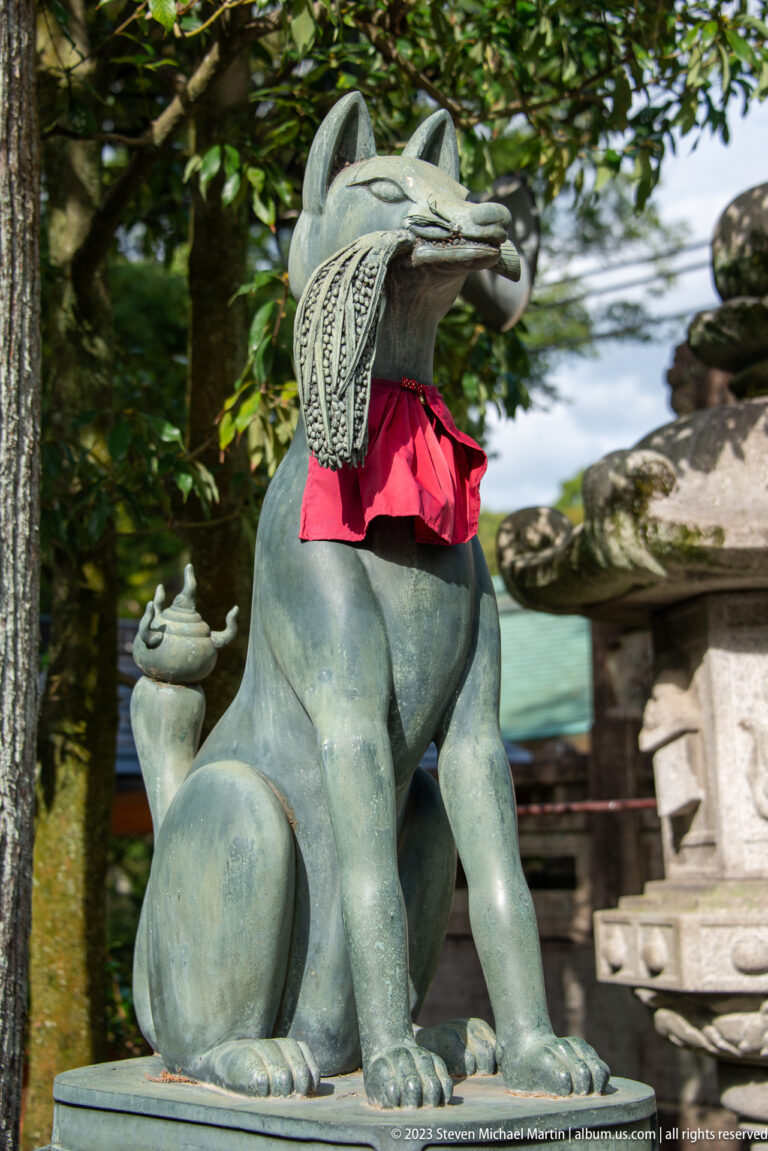 Seated bronze fox guardian wearing a red votive bib beneath trees.
