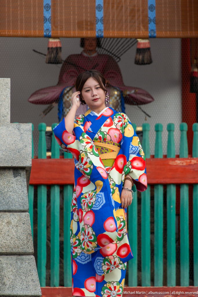Visitor in a bright blue floral kimono posing beside a vermilion and green railing at Fushimi Inari Taisha.