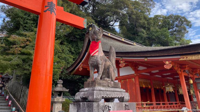 Stone fox guardian with a red bib beside a vermilion torii and shrine.
