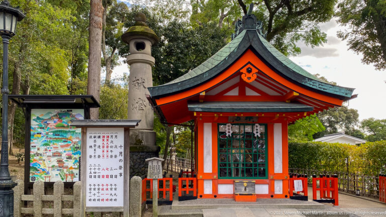 Small vermilion subsidiary shrine with green roof beside a stone monument and map board.