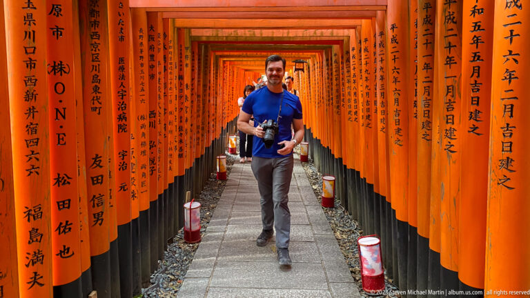 Photographer walking through an inscribed vermilion torii corridor.