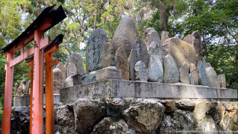 Cluster of carved stone memorial monuments beside a vermilion torii in a wooded shrine setting.
