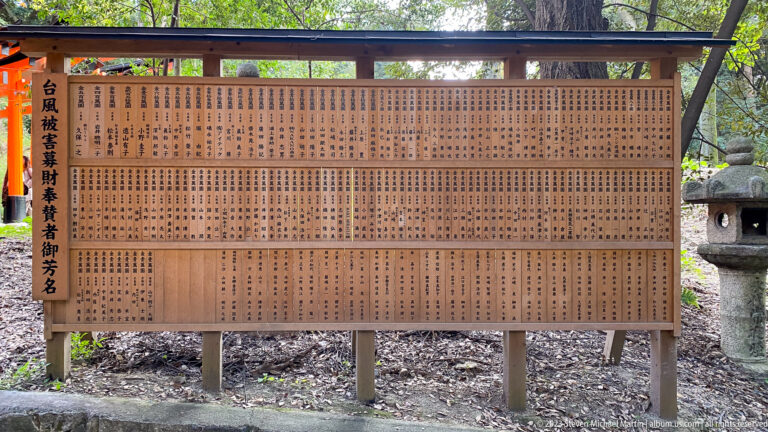 Large wooden board covered with vertical Japanese inscriptions beside a shrine path.