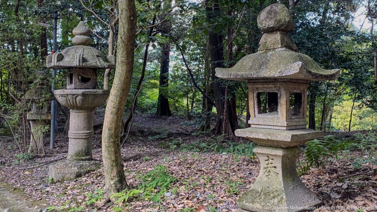 Two traditional carved stone lanterns standing in a wooded shrine setting.