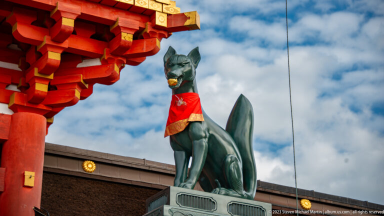 Seated bronze fox guardian with a red votive bib beside vermilion shrine architecture under a cloudy sky.