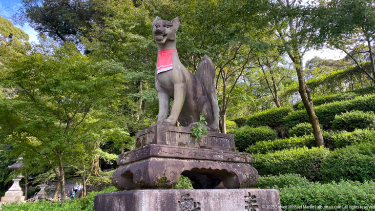 Stone kitsune guardian wearing a red votive bib on a pedestal among trees.