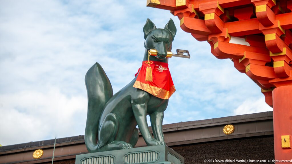 Bronze fox guardian holding a key in its mouth, wearing a red bib beneath vermilion shrine brackets.